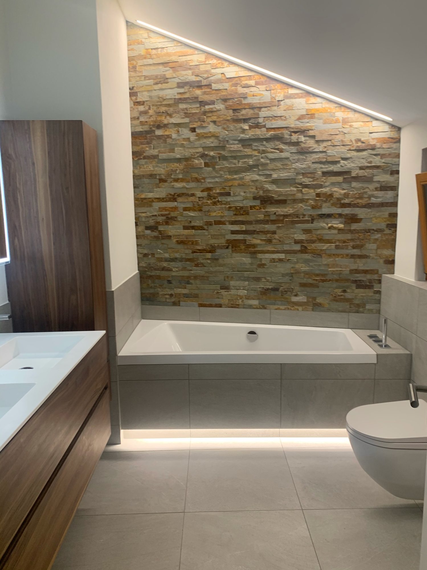 Saniertes Dachbad mit Natursteinwand, indirekter LED-Beleuchtung, Einbauwanne und Nussbaum-Möbel